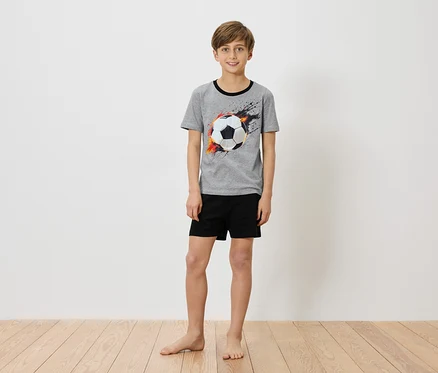Chlapec stojí bosý pred bielou stenou a má na sebe sivé tričko s potlačou futbalovej lopty a čierne šortky.