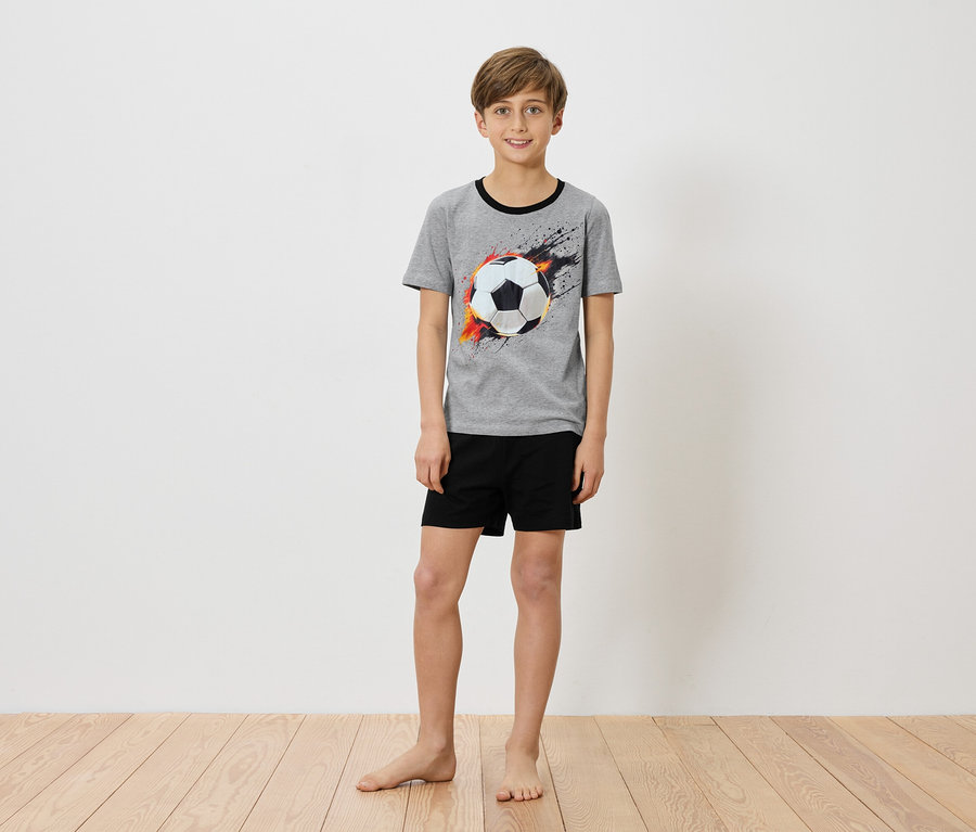 Chlapec stojí bosý pred bielou stenou a má na sebe sivé tričko s potlačou futbalovej lopty a čierne šortky.