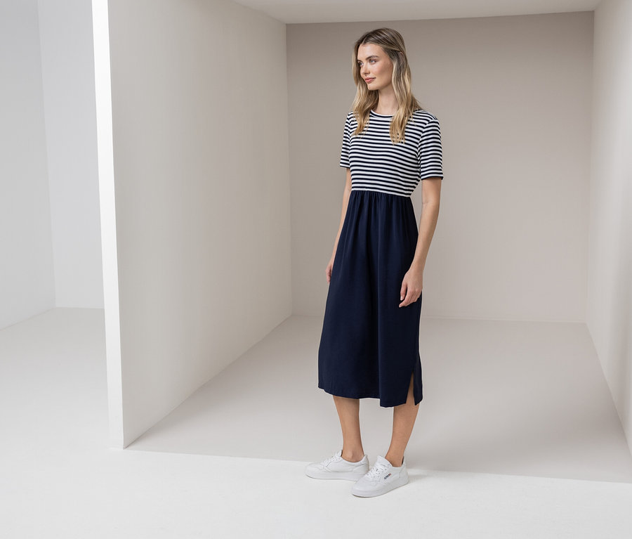 Žena stojí v bielej miestnosti a má na sebe modro-biele pruhované šaty a biele tenisky.