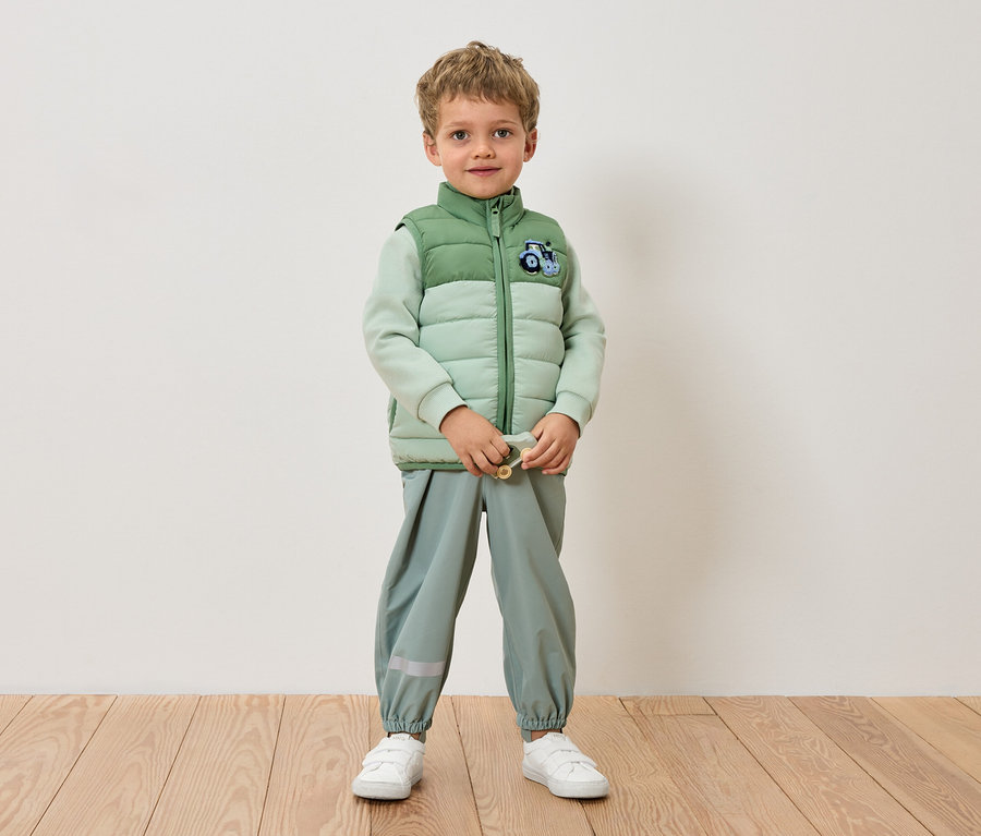 Chlapec stojí pred stenou v svetlozelenej bunde, svetlozelených nohaviciach a bielych teniskách.