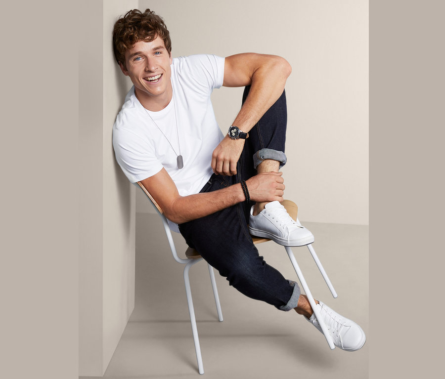 Muž sedí na stoličke a má na sebe biele tričko z 2 ks sady tričiek s okrúhlym výstrihom, džínsy a tenisky.