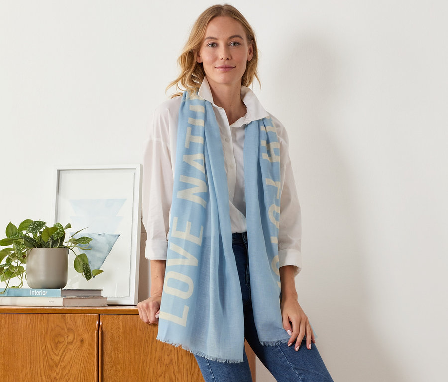 Žena sa opiera o skrinku a má na sebe tkaný šál so statement potlačou.