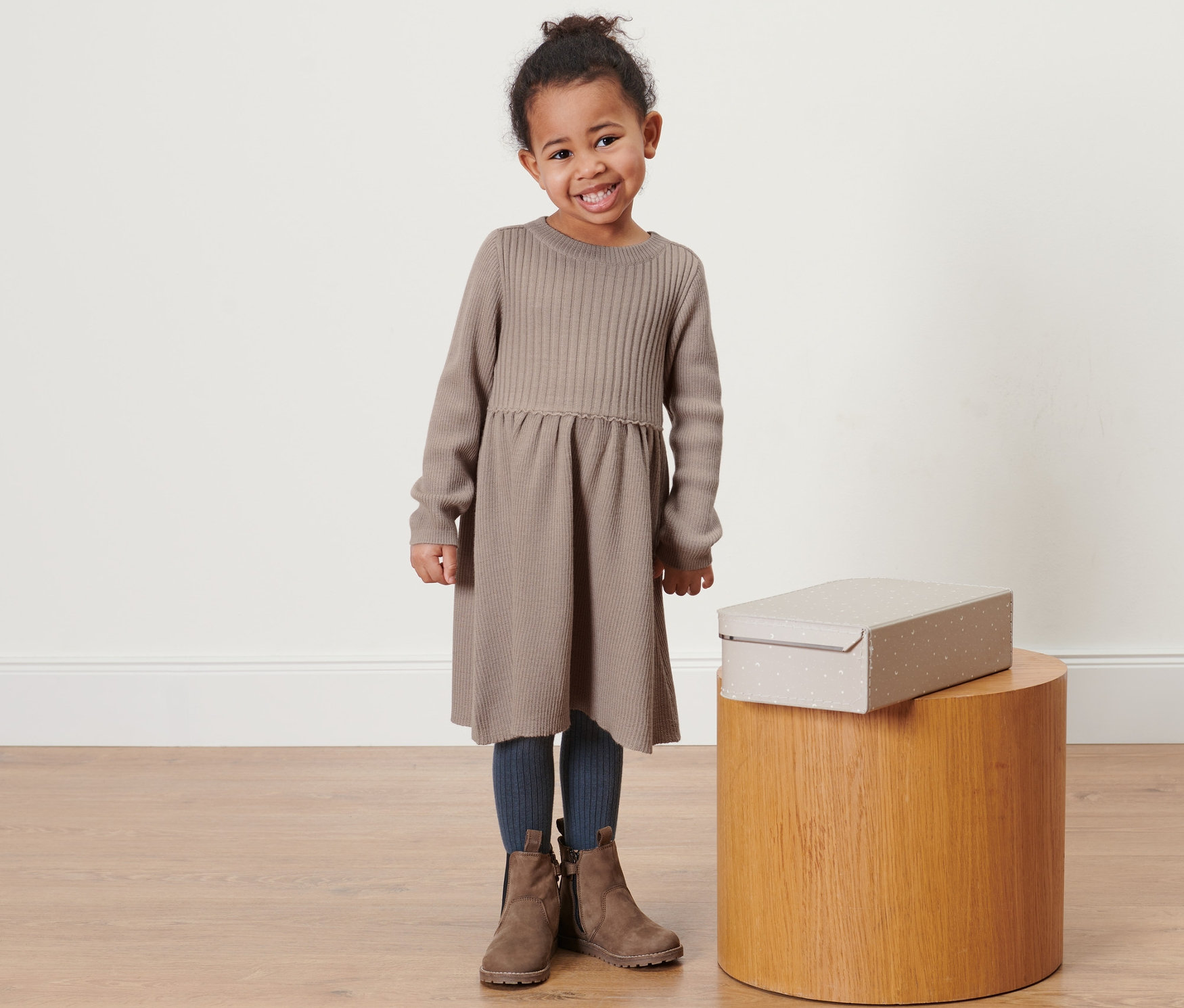 Usmievajúce sa dievčatko pózuje v pletených šatách s merino vlnou svetlohnedej farby. Vedľa nej stojí na drevenej stoličke krabica.