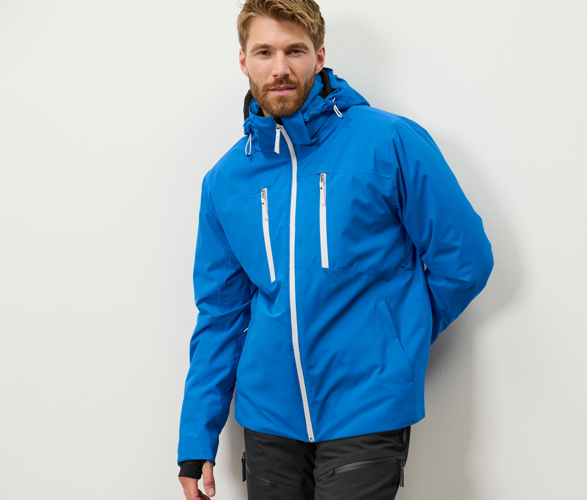 Muž s bradou pózuje v modrej high-tech lyžiarskej bundě.