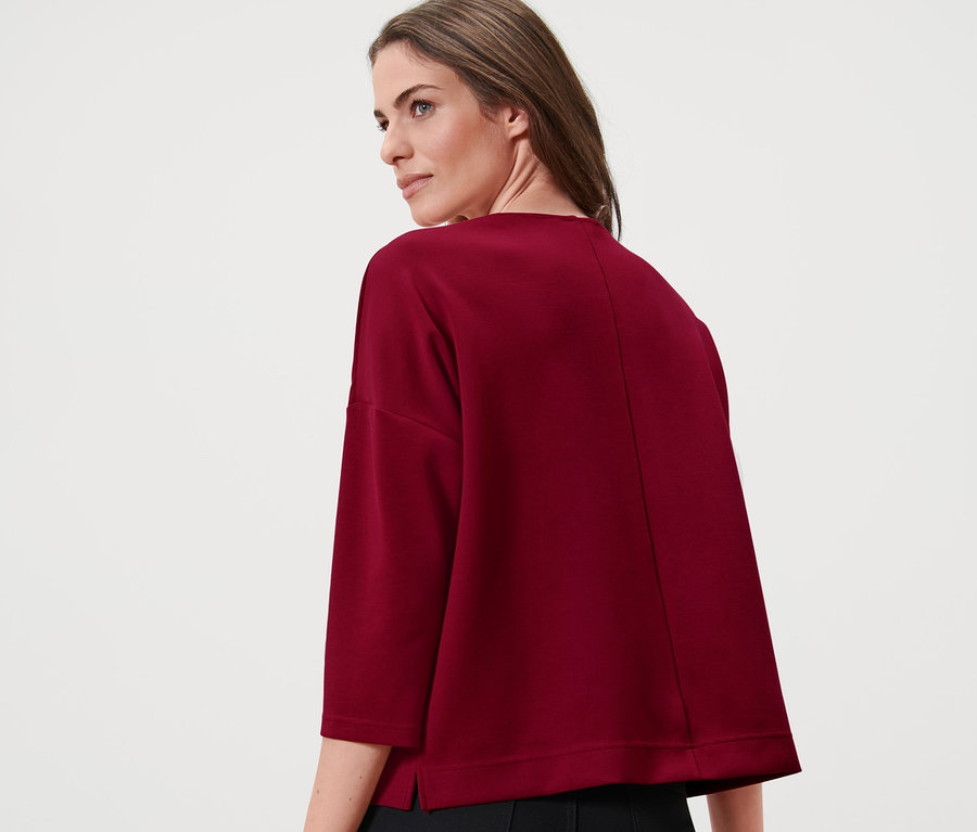 Žena má na sebe bordové mikinové tričko s 3/4 rukávmi a čierne nohavice.