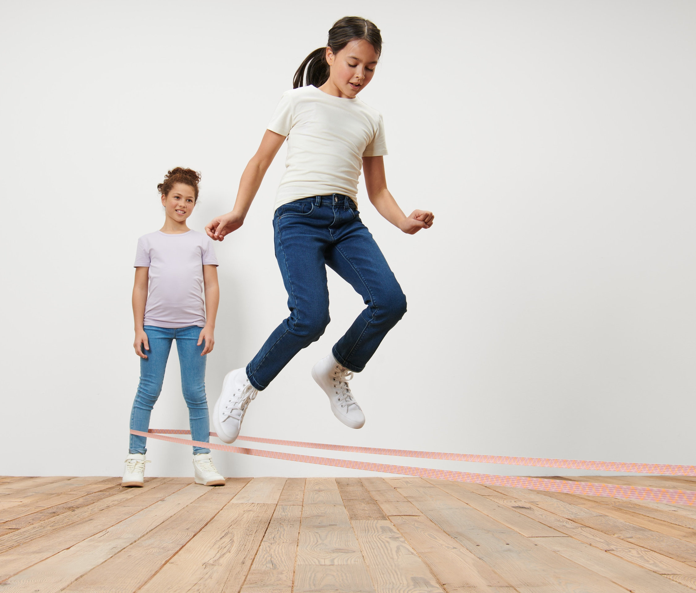 Dievča skáče cez gumu na skákanie, druhé dievča gumu drží.