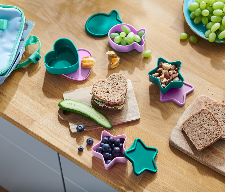 Na drevenom stole je snack. Srdcový sendvič, uhorka, čučoriedky v nádobe v tvare hviezdy, orechy, hrozno v nádobe v tvare ryby a chlieb.