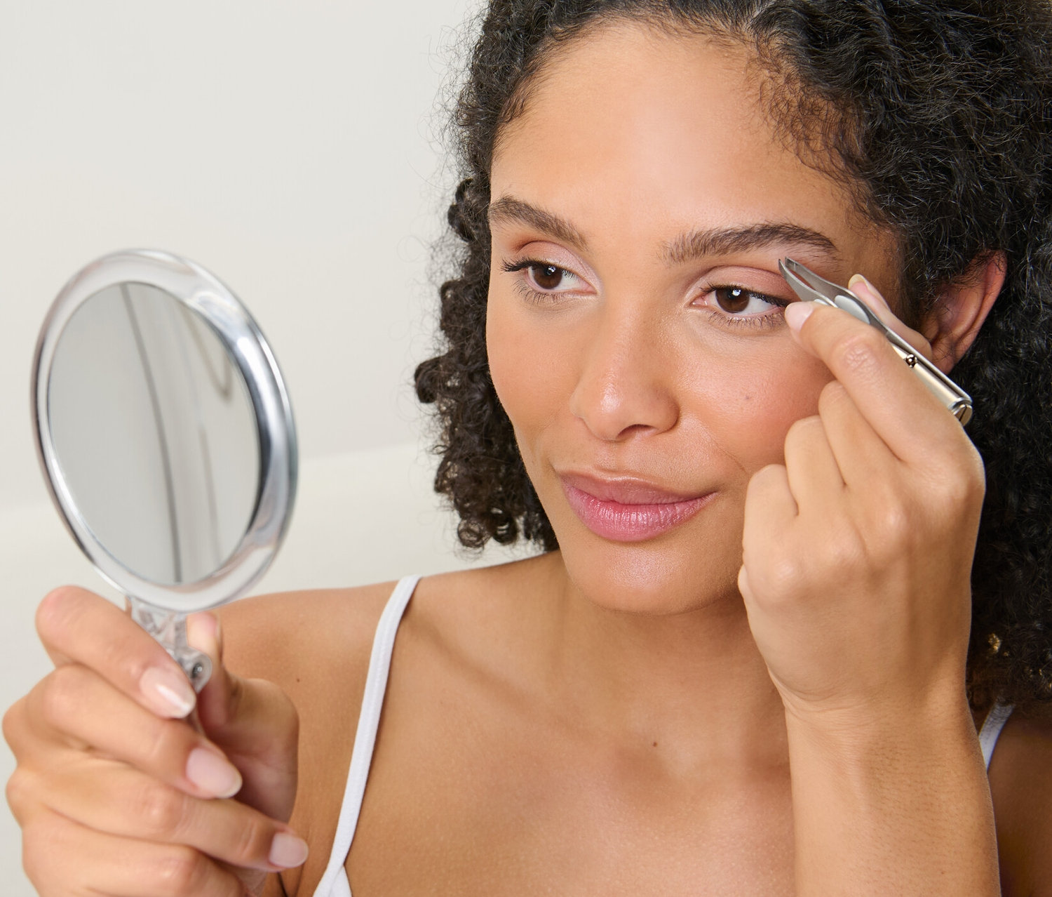 Žena si pinzetou s LED svetlom upravuje obočie a zároveň sa pozerá do zrkadla.