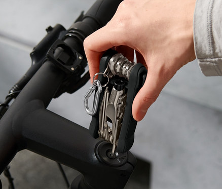 Ruka drží multifunkčný nástroj na bicykel na riadidlách bicykla. V pozadí je vidieť helmu.