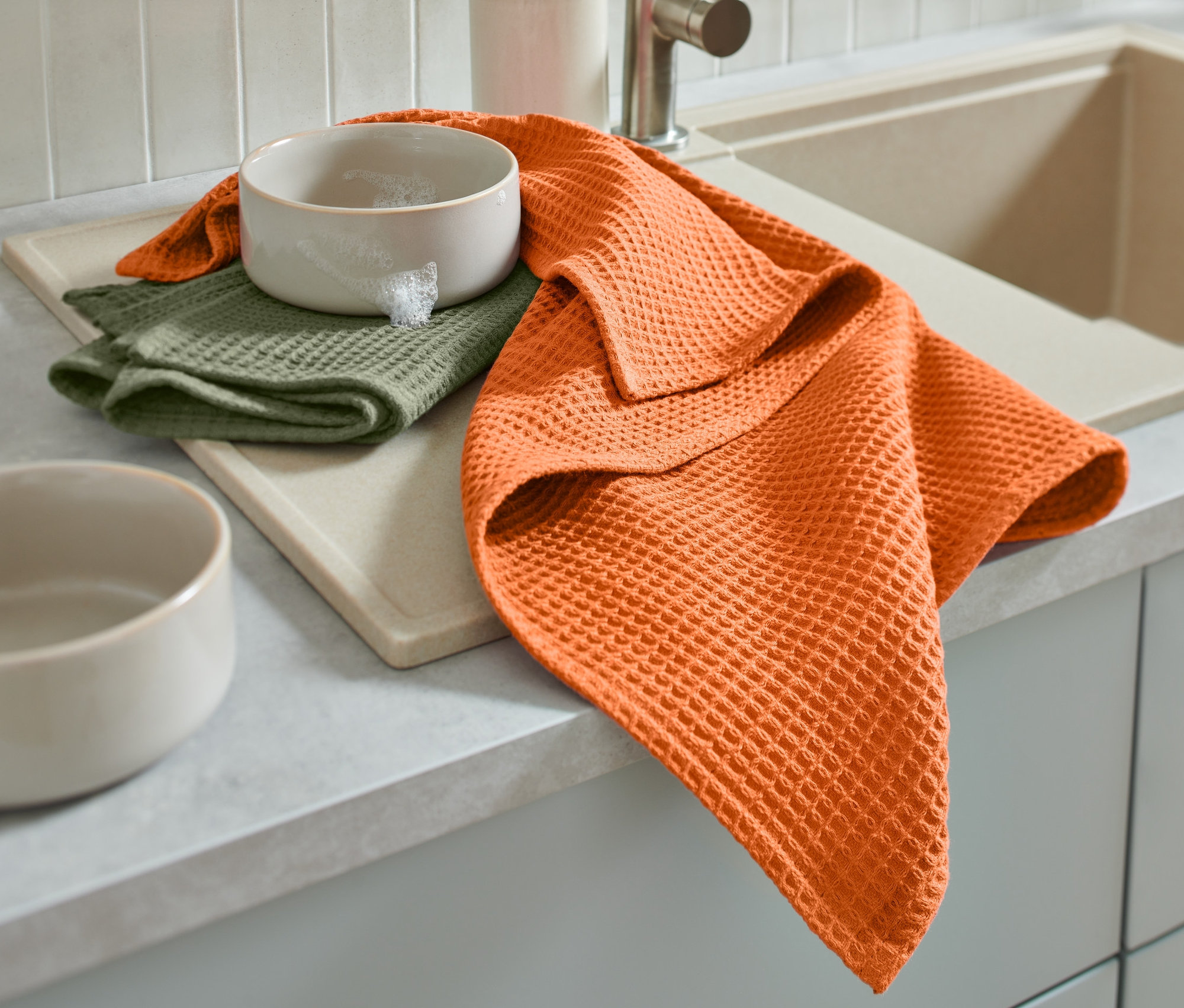 Oranžová utierka a zložená zelená utierka ležia vedľa misky s penou na doske pri dreze.