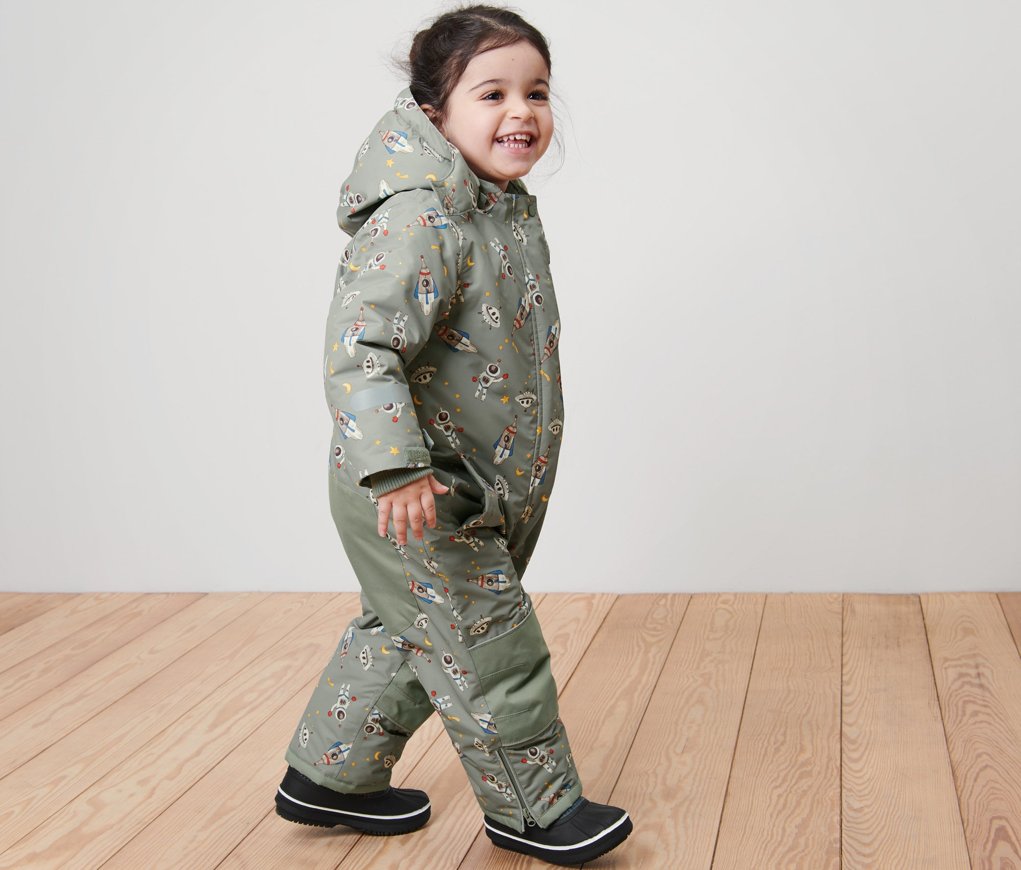 Usmievajúce sa dieťa kráča a má na sebe detskú zimnú kombinézu s potlačou vesmíru.