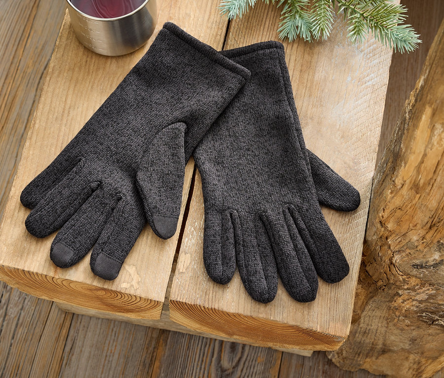 Pár sivých rukavíc leží na drevenom povrchu vedľa strieborného hrnčeka a vetvičky ihličnanu.