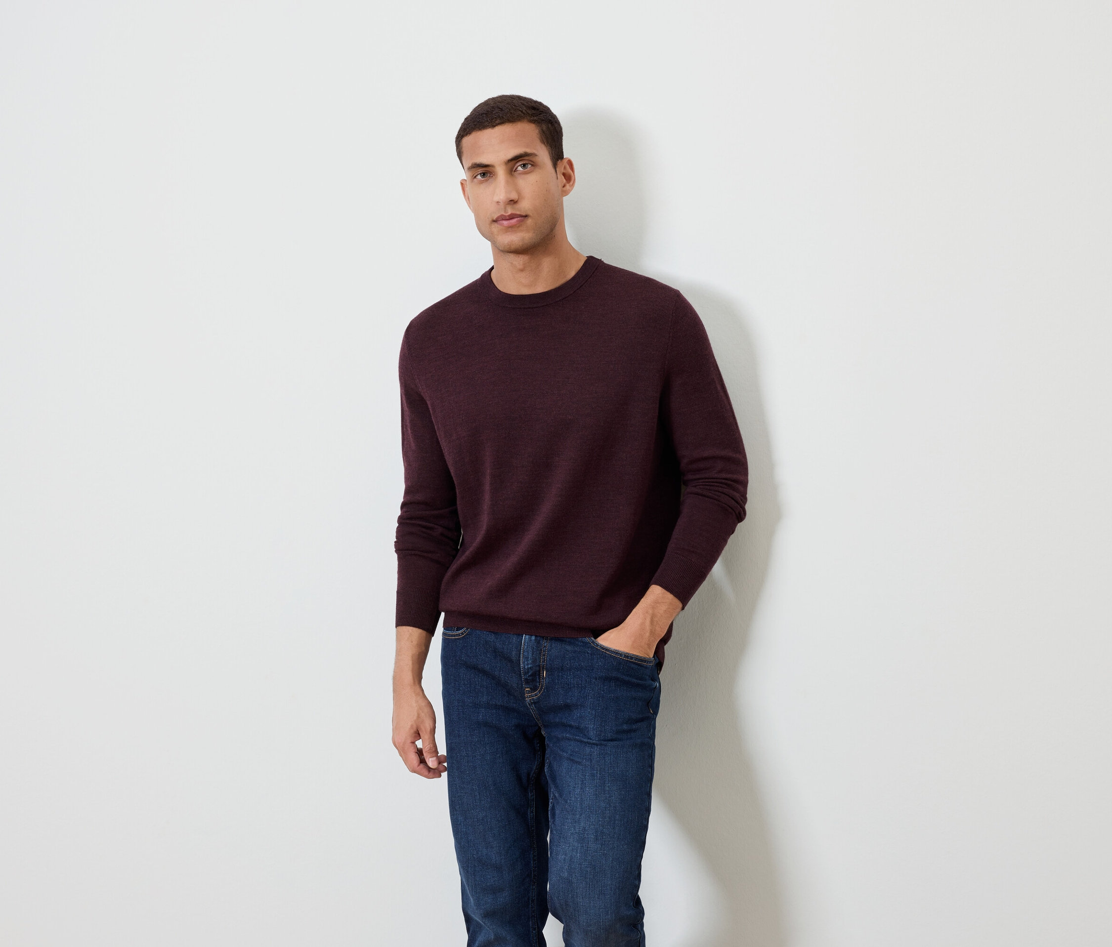 Muž pózuje na bielom pozadí a má na sebe bordový sveter a džínsy.