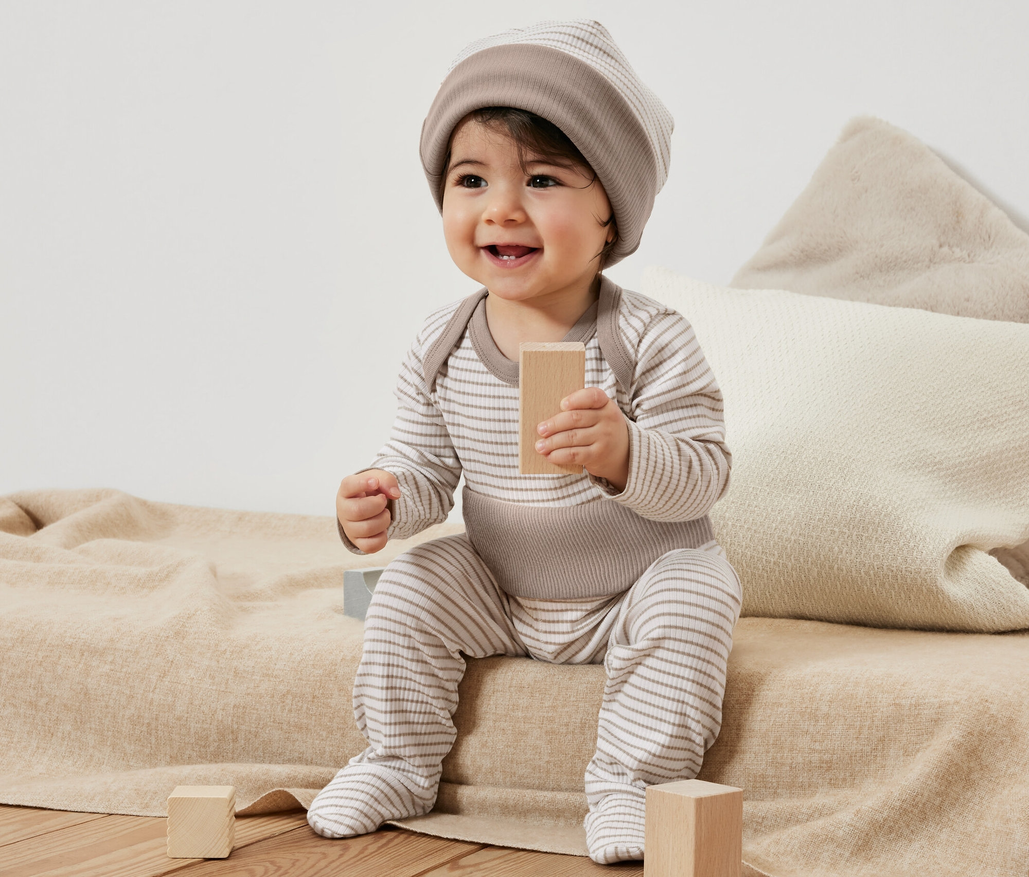 Usmievajúce sa dieťa sedí na deke a hrá sa s drevenými kockami. Má na sebe pruhovanú dojčenskú súpravu, 3-dielnu.