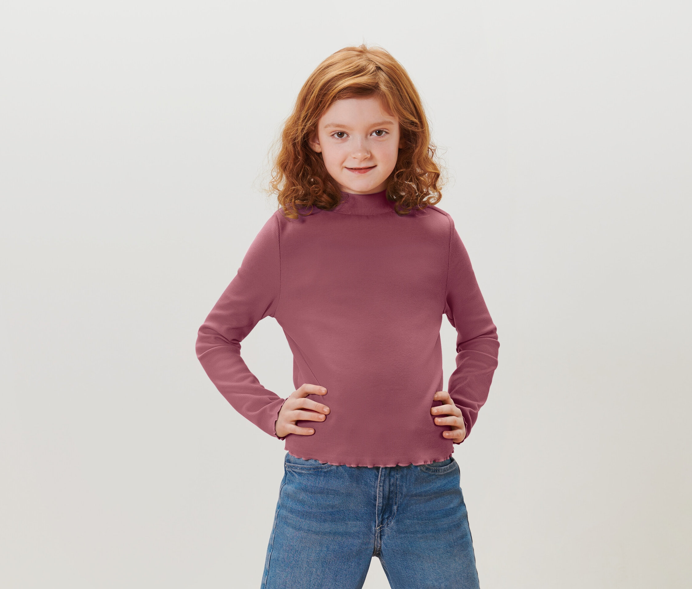 Dievča s ryšavými kučeravými vlasmi stojí s rukami v bok. Má na sebe purpurové tričko s dlhým rukávom a modré džínsy.