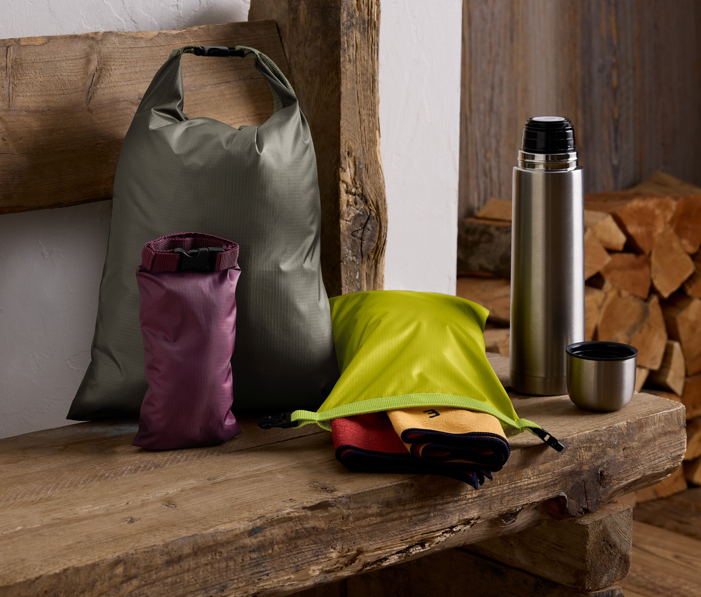Na drevenej lavici leží zelený vak, fialový vak, zelený vak s uterákmi, termoska a stoh dreva.