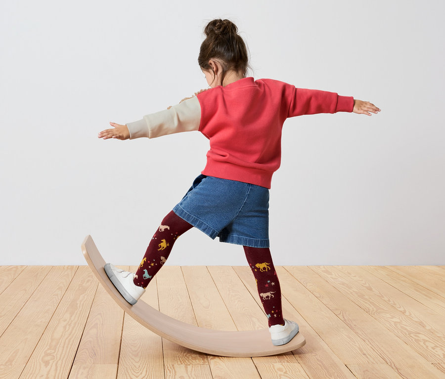 Dievča stojí na balančnej doske, má na sebe červeno-krémovú mikinu, džínsové kraťasy a bordové pančuchy s koňmi.