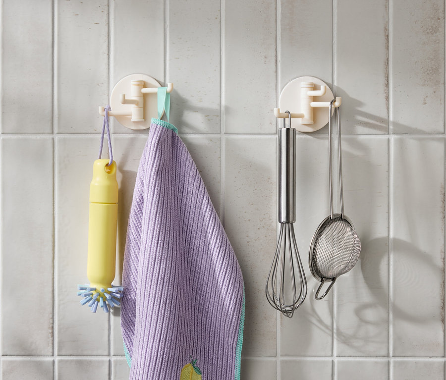Na bielych kachličkách visí žltá kefa, fialová utierka, nerezová metla a sitko.