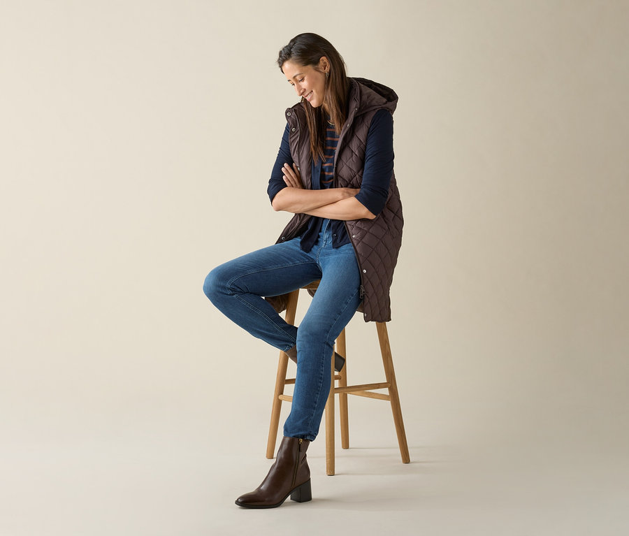 Žena sedí na stoličke so skríženými rukami na hrudi a má na sebe dlhú prešívanú vestu s kapucňou v tmavohnedej farbe.