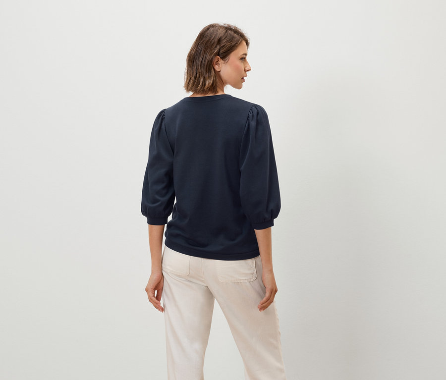 Žena má na sebe tmavomodrý top s trojštvrťovými rukávmi a béžové nohavice.