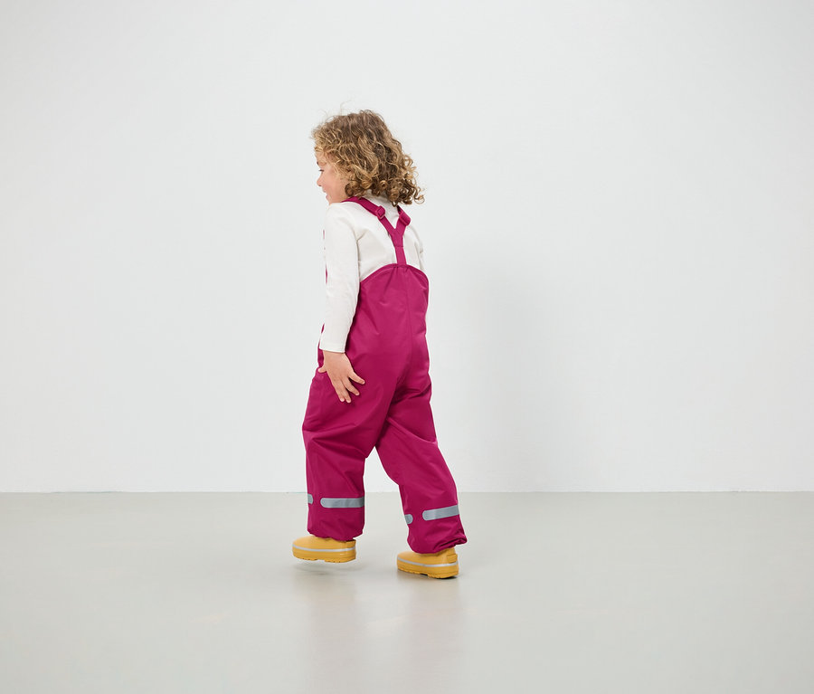 Dievča v ružových nohaviciach s náprsenkou a žltých topánkach kráča po podlahe.