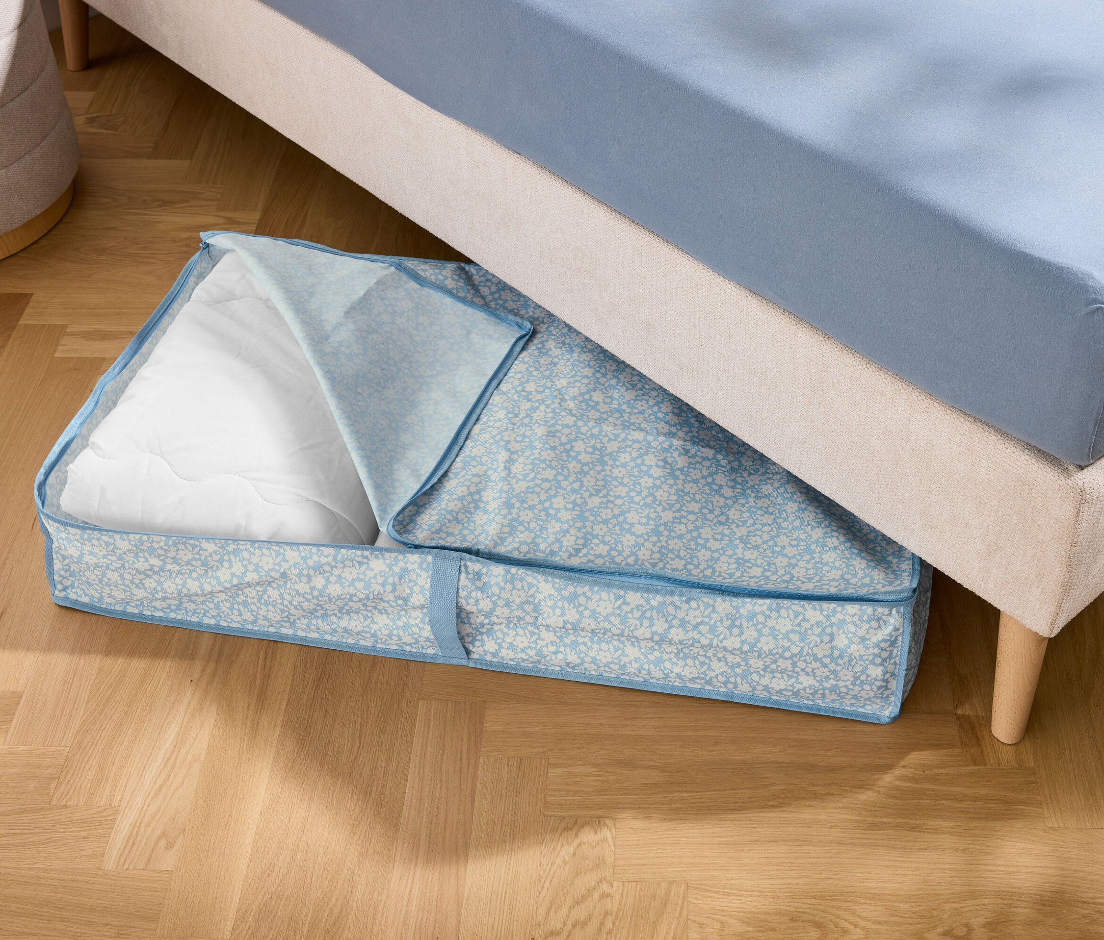 Pod posteľou leží modrá úložná taška s bielou výplňou.