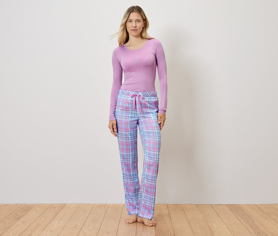 Žena v lila tope a kockovaných pyžamových nohaviciach stojí na drevenej podlahe.