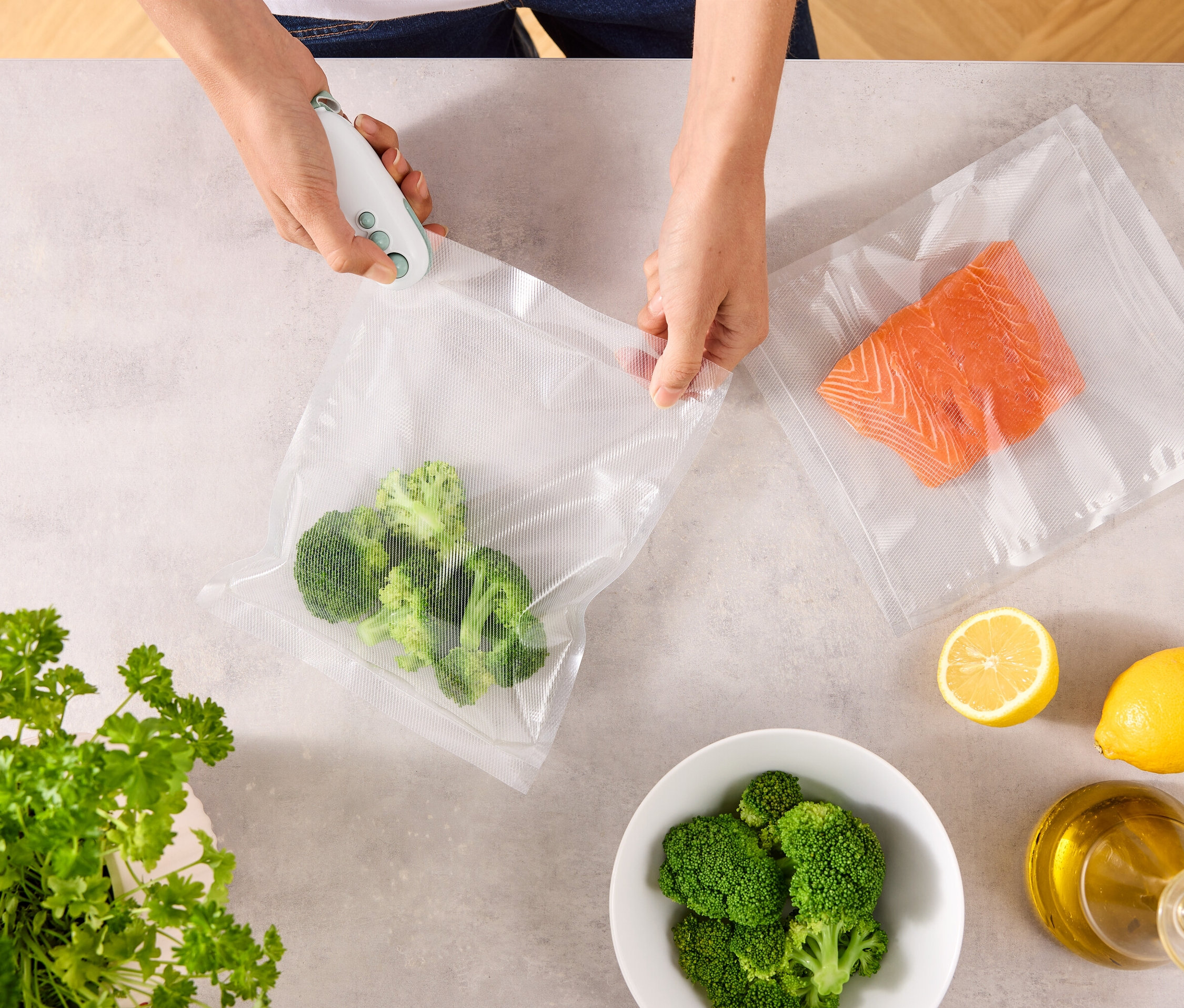 Osoba uzatvára vrecko s brokolicou pomocou vákuovačky. Na stole leží ďalšie vrecko s lososom, citróny, miska brokolice, petržlen a fľaša oleja.