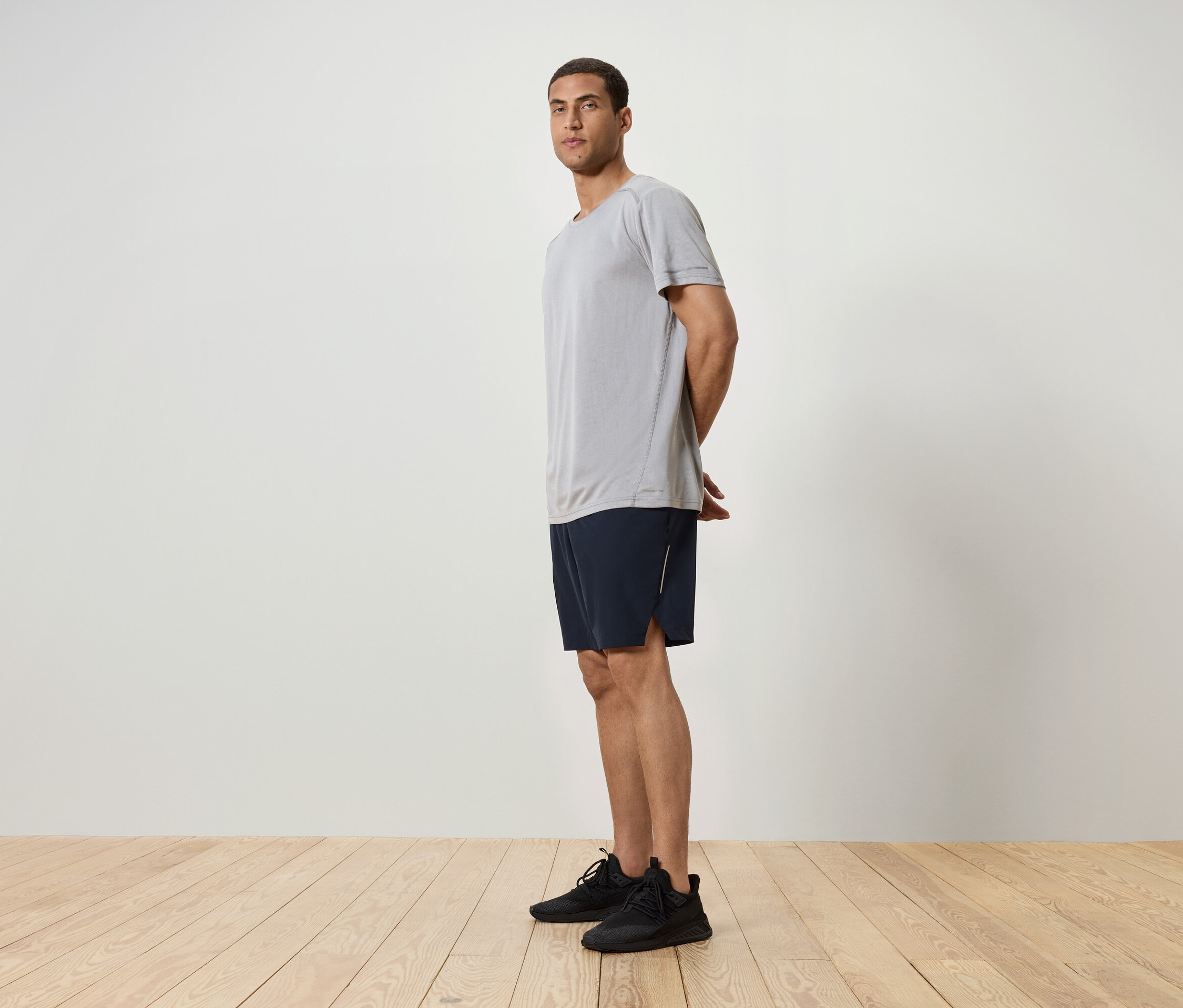 Muž stojí bokom, má na sebe sivé tričko, tmavomodré šortky a čierne tenisky.