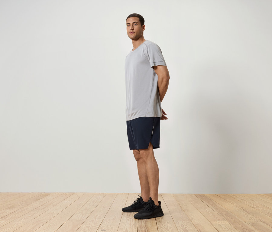 Muž stojí bokom, má na sebe sivé tričko, tmavomodré šortky a čierne tenisky.