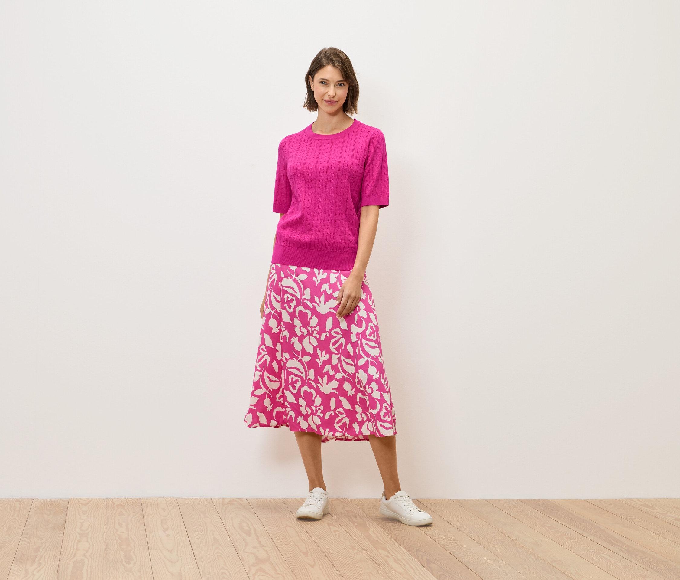 Žena v ružovom svetri a sukni s kvetinovým vzorom stojí na drevenej podlahe.