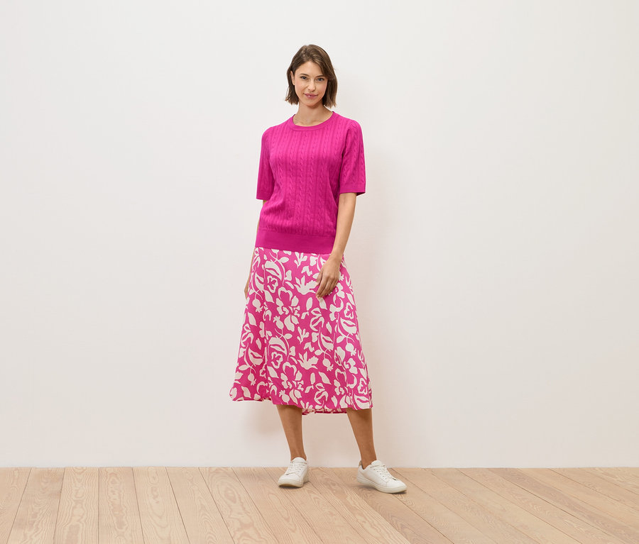 Žena v ružovom svetri a sukni s kvetinovým vzorom stojí na drevenej podlahe.