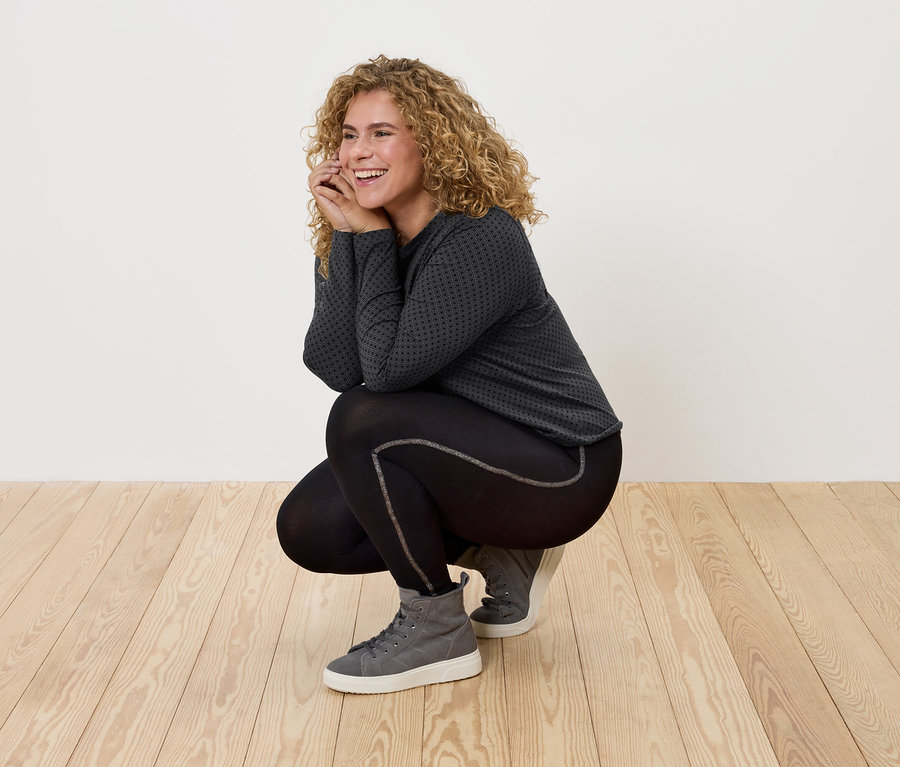 Žena s kučeravými vlasmi v čiernom športovom oblečení a sivých teniskách pózuje na drevenej podlahe.
