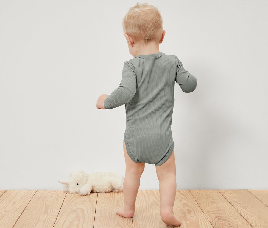 Malé dieťa v rebrovanom body s dlhým rukávom stojí na drevenej podlahe vedľa plyšového zvieraťa.