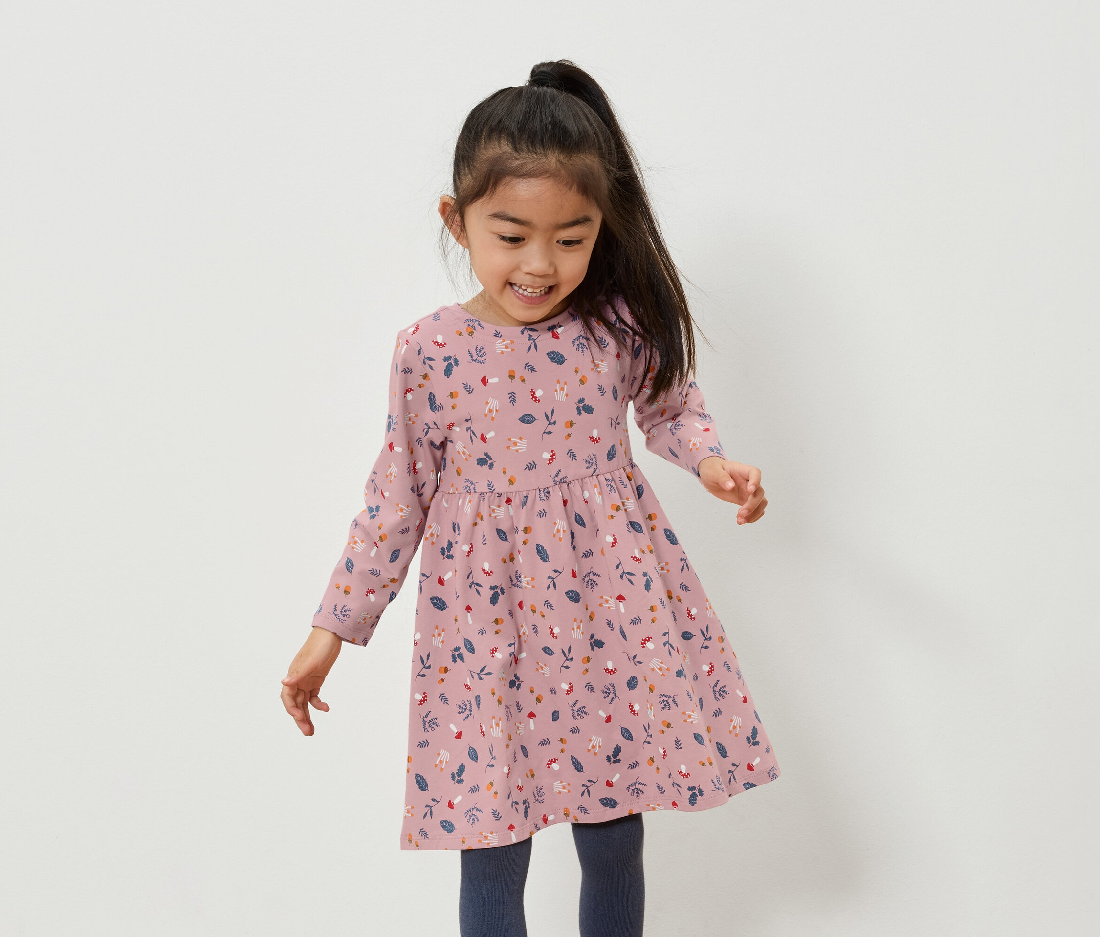 Usmievajúce sa ázijské dievča v ružových šatách s kvetinovou potlačou a modrých legínach.