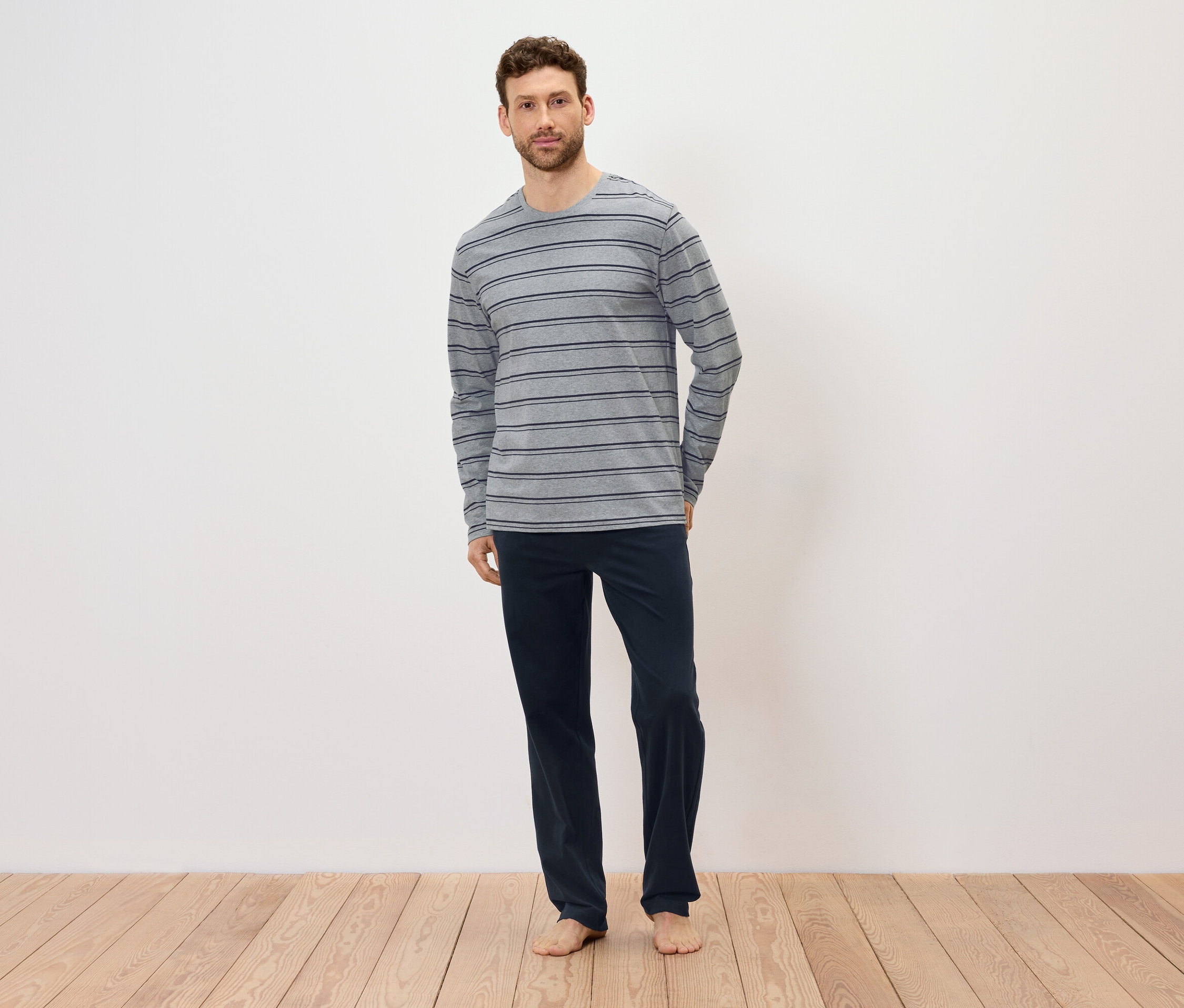 Muž stojí naboso na drevenej podlahe, má na sebe sivé pruhované tričko s dlhým rukávom a tmavomodré nohavice.