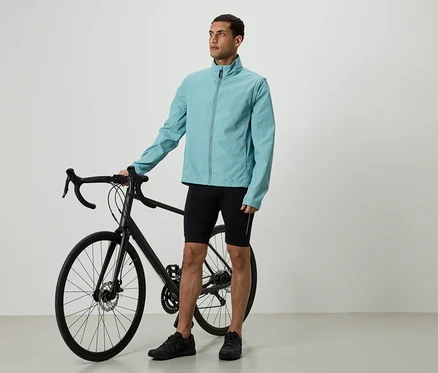 Muž v svetlomodrej bunde a čiernych šortkách stojí vedľa čierneho bicykla.