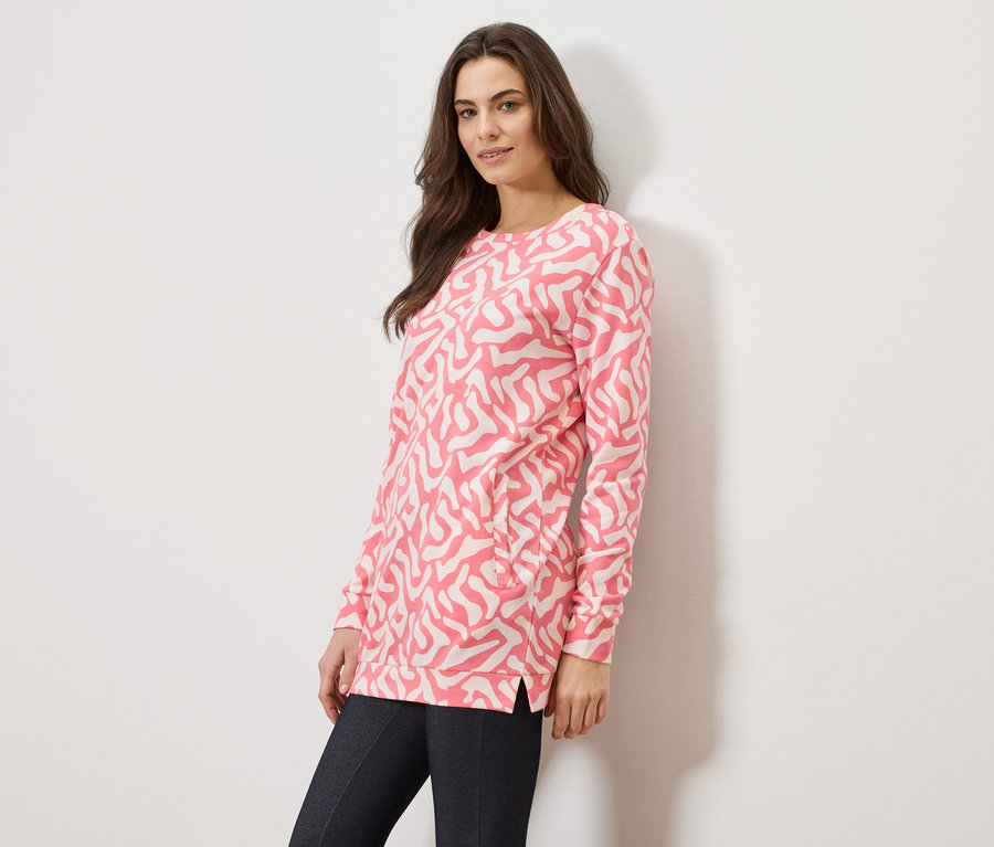 Žena má na sebe ružový sveter s abstraktným vzorom a tmavé džínsy na bielom pozadí.