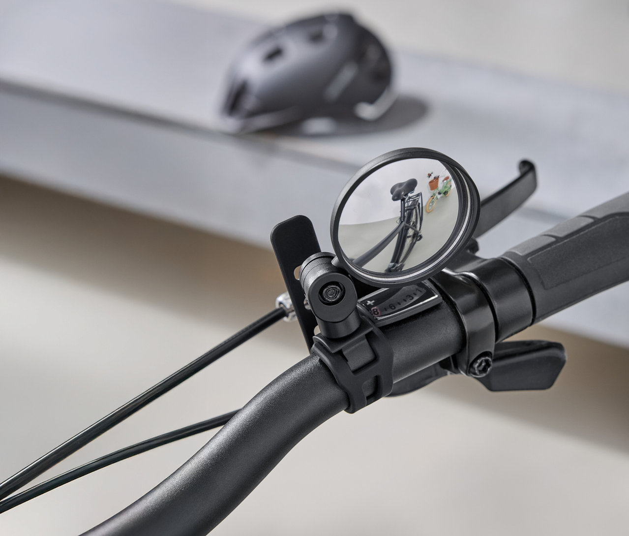Detailný záber na riadidlá bicykla s okrúhlym spätným zrkadlom, ktoré odráža cestu za bicyklom. V pozadí je sivá cyklistická prilba.
