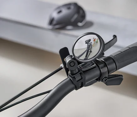 Detailný záber na riadidlá bicykla s okrúhlym spätným zrkadlom, ktoré odráža cestu za bicyklom. V pozadí je sivá cyklistická prilba.