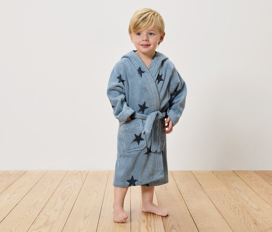 Malý chlapec s blond vlasmi stojí naboso na drevenej podlahe a má na sebe svetlomodrý župan s tmavými hviezdami.