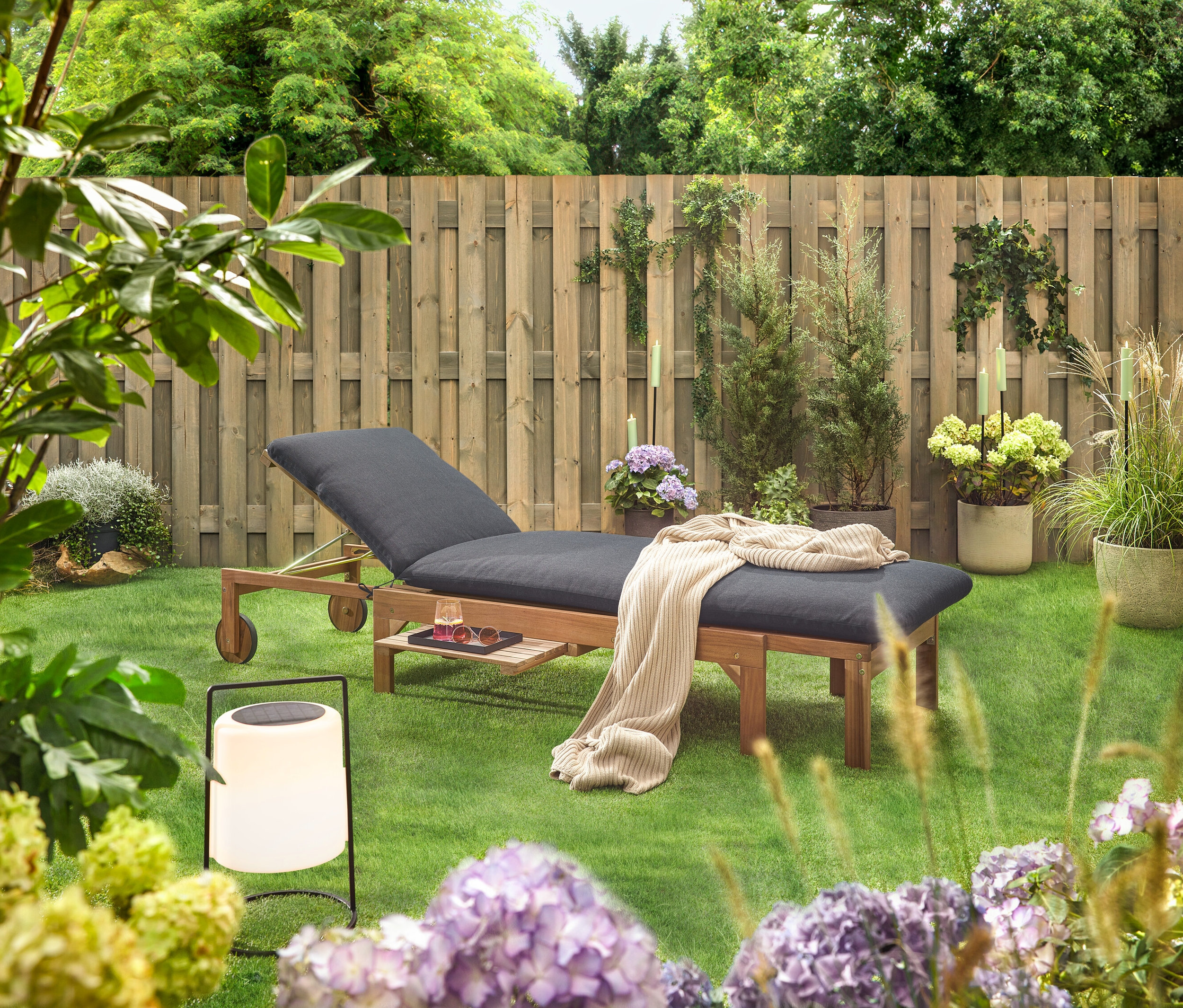 Záhradná scéna s dreveným ležadlom s čiernym čalúnením, dekou, nápojom, slnečnými okuliarmi a lampášom na trávniku pred dreveným plotom.