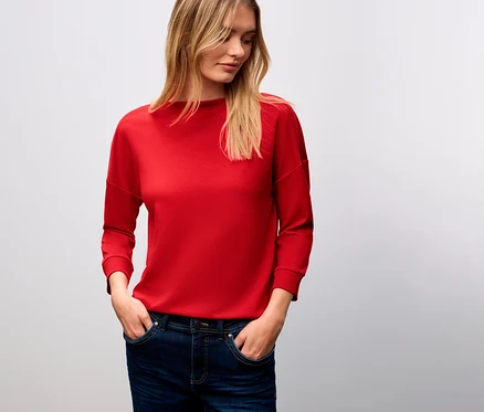 Žena s blond vlasmi má na sebe červené tričko Street One s rebrovanými prvkami a džínsy.