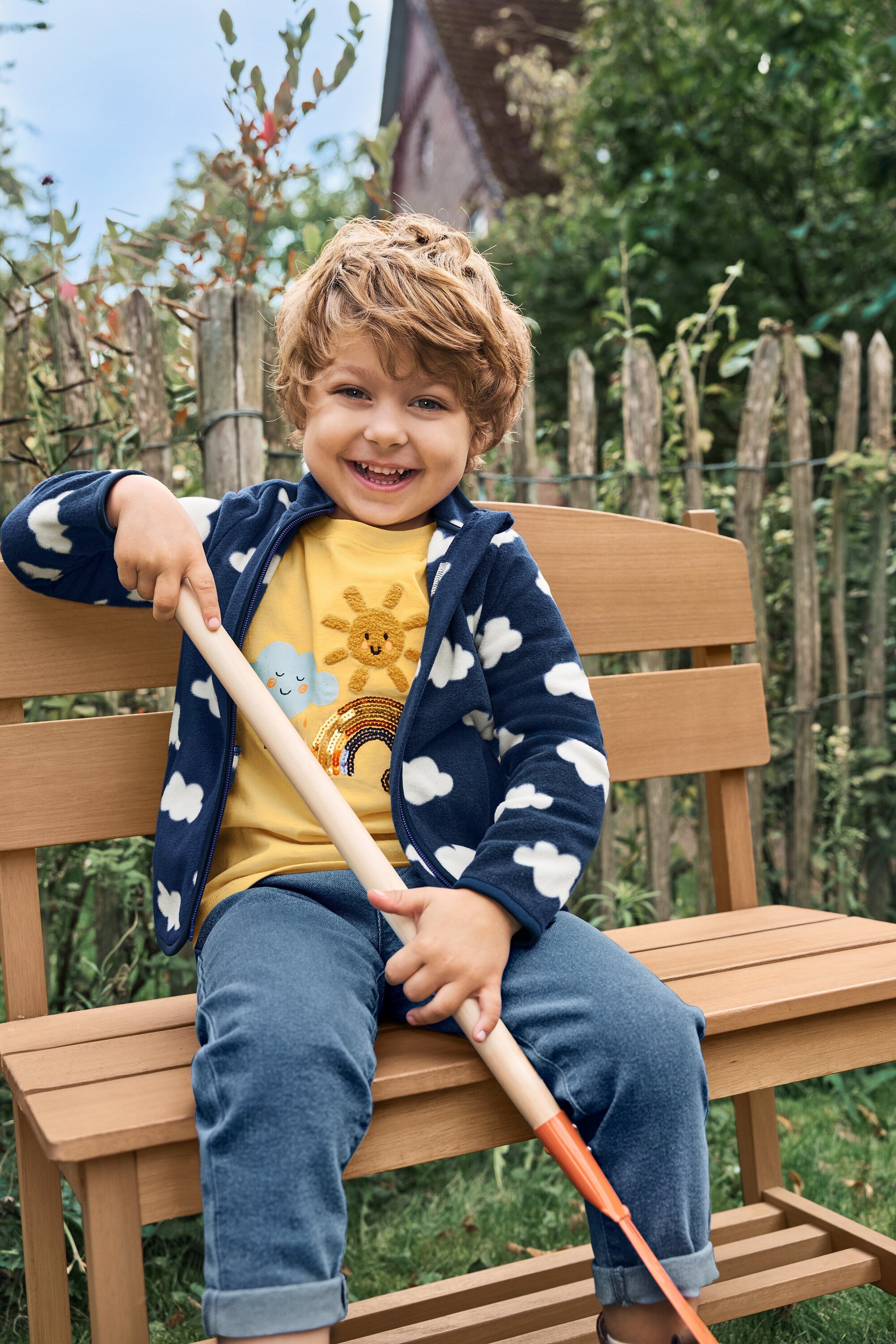 Usmievavý chlapec sedí na malej záhradnej lavičke s odkladacím priestorom a drží lopatu.