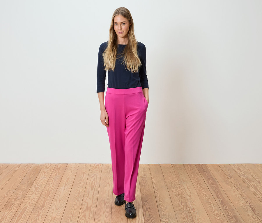 Žena stojí v tmavomodrej blúzke a ružových nohaviciach pred bielou stenou.