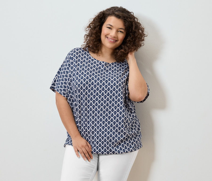 Žena s kučeravými vlasmi v modro-bielej blúzke a bielych nohaviciach sa usmieva.