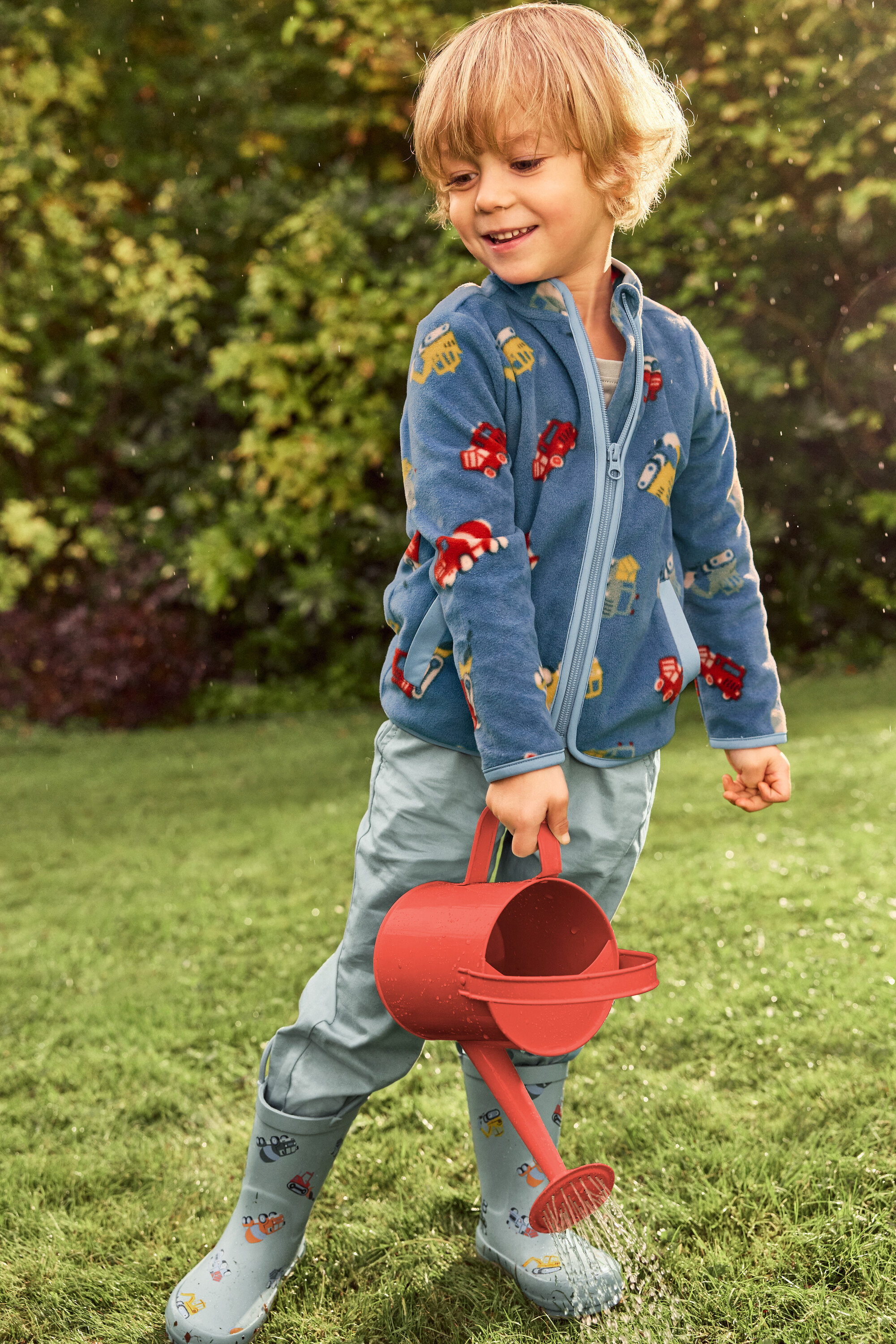 Usmievavý chlapec s blond vlasmi polieva červenou kanvou zelený trávnik. Má na sebe modrú bundu s motívmi vozidiel a sivé gumáky s vozidlami.