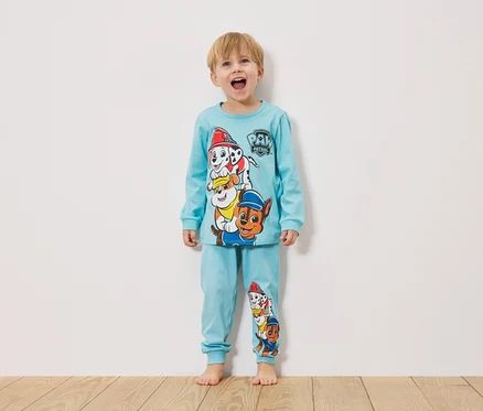 Malý chlapec stojí pred bielou stenou a má na sebe svetlomodré pyžamo Paw Patrol.