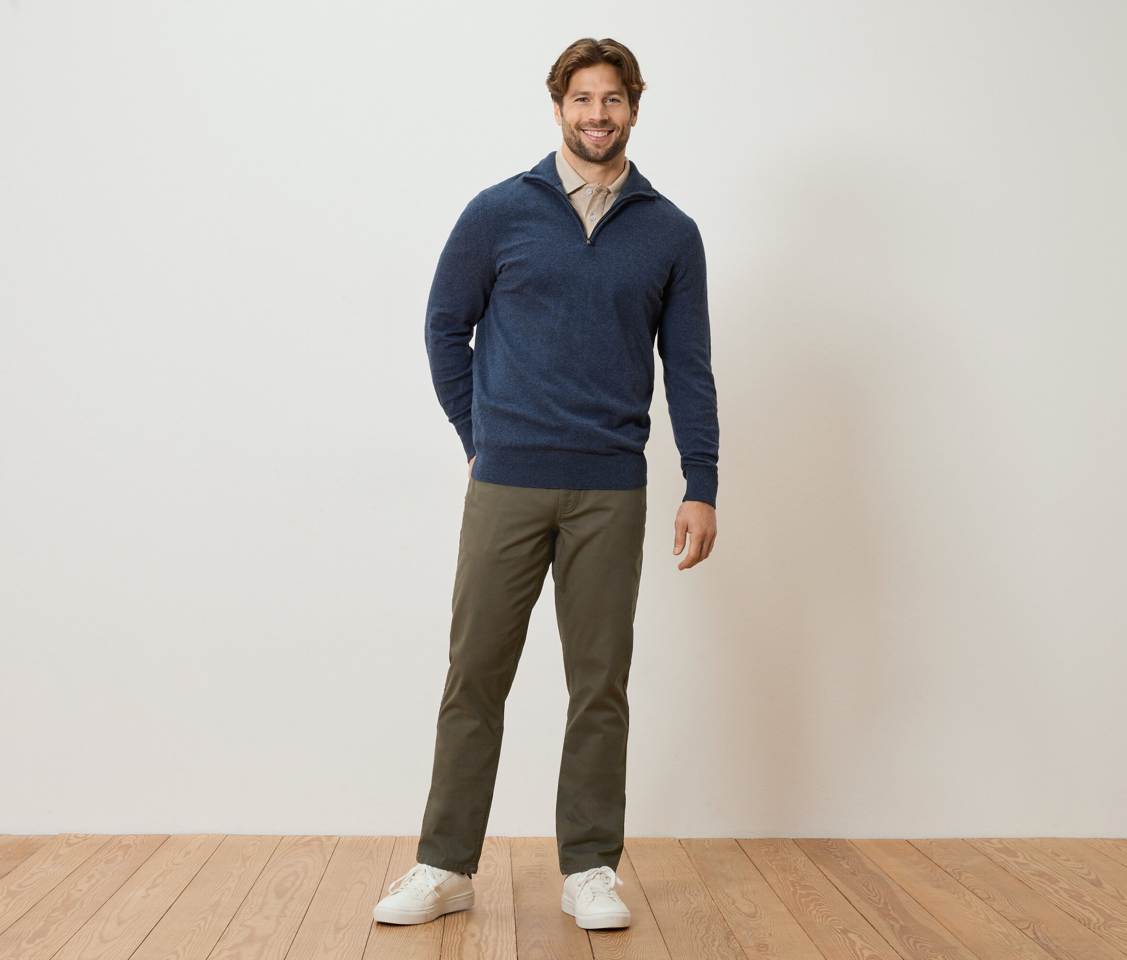 Muž stojí s rukami vo vreckách pred stenou. Má na sebe tmavomodrý sveter, svetlohnedú blúzku, khaki nohavice a biele tenisky.