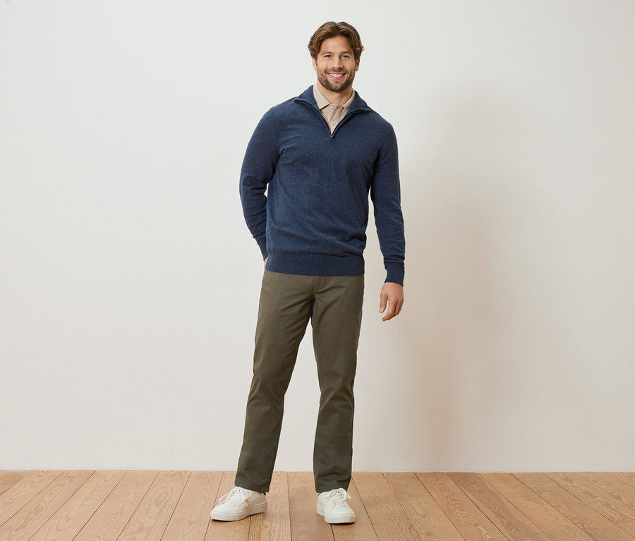 Muž stojí s rukami vo vreckách pred stenou. Má na sebe tmavomodrý sveter, svetlohnedú blúzku, khaki nohavice a biele tenisky.